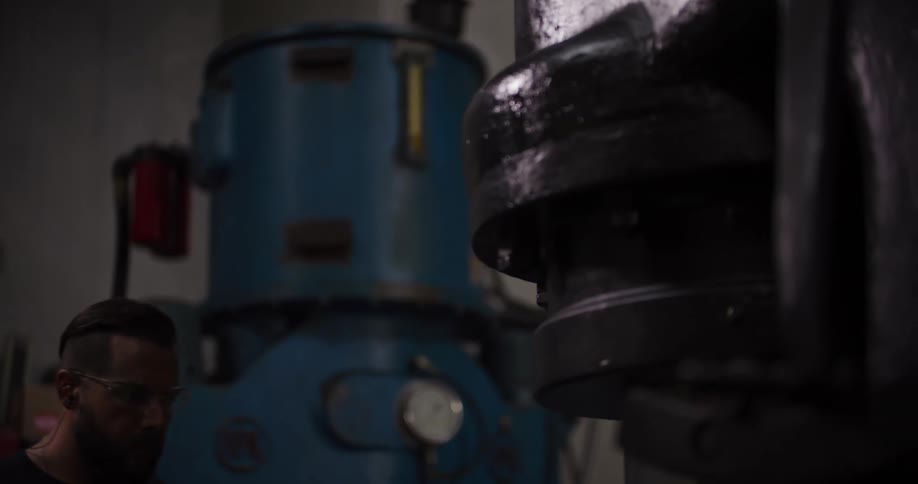 A strong, tattooed blacksmith in a dark workshop carefully operates a large industrial press in slow motion