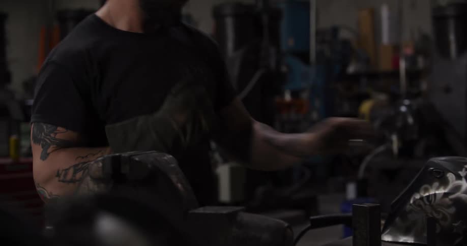 A tattooed and bearded metalworker puts on his protective gloves and welding helmet in a dimly lit workshop