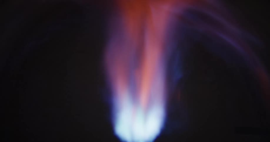 A macro slow-motion view shows a gas burner igniting, with the blue flame dancing and turning orange against a dark background