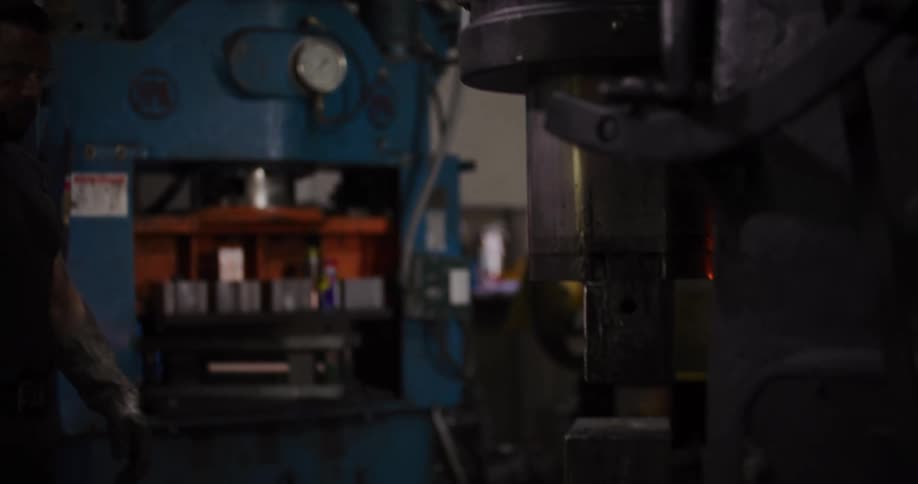 An industrial worker with safety gear carefully adjusts a large power hammer machine before walking away as it operates