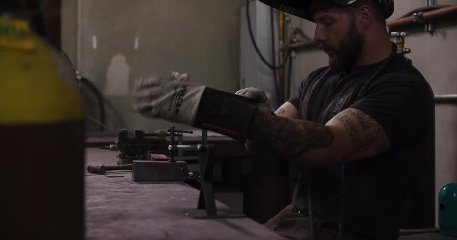 A tattooed craftsman puts on his welding helmet and protective gloves before TIG welding a piece of metal in his workshop