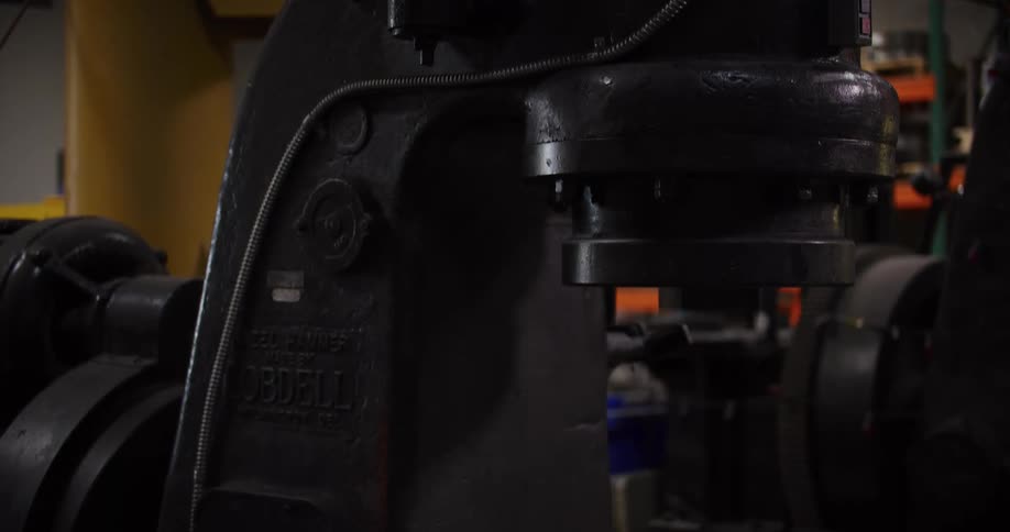A worker activates a massive vintage industrial trip hammer in a dark factory setting