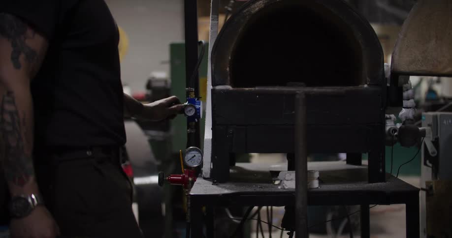 A craftsman with tattooed arms uses a blowtorch to ignite a gas-powered forge in a metalworking workshop