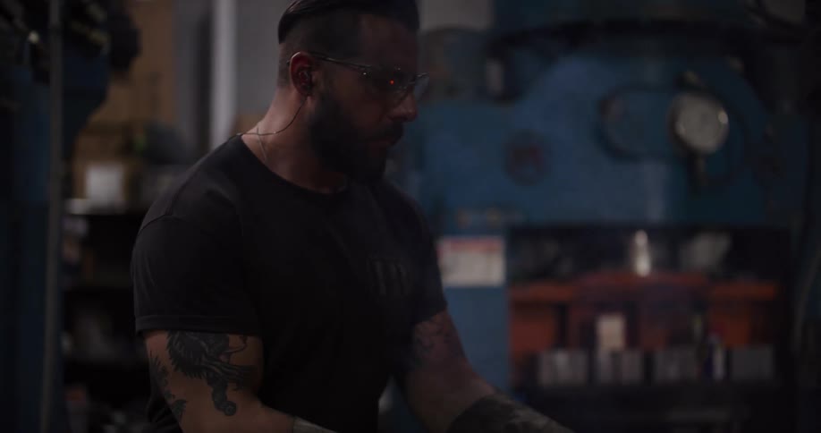 A focused blacksmith wearing safety glasses works in a smoky industrial forge as the camera slowly pulls back