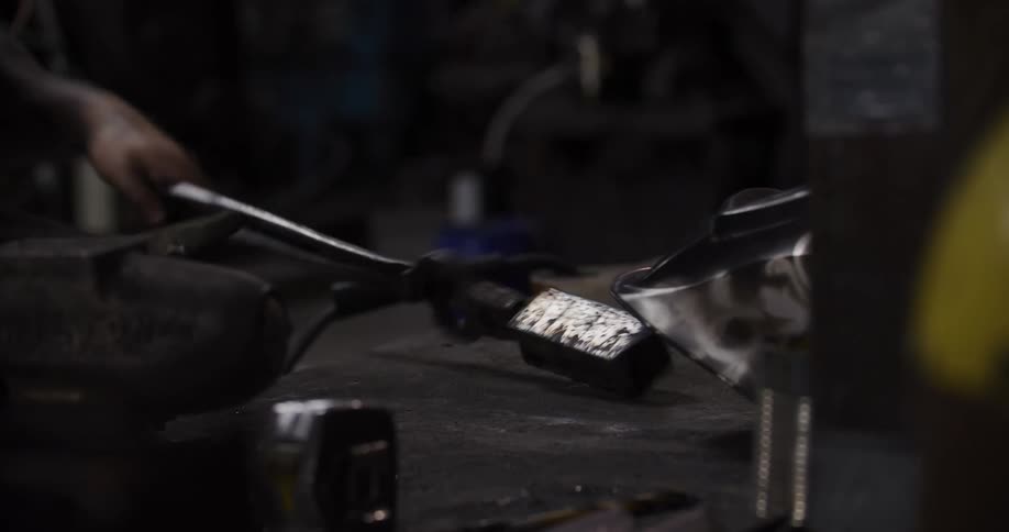 A tattooed metalworker in a dimly lit workshop puts down his welding helmet and tools on a workbench before walking away
