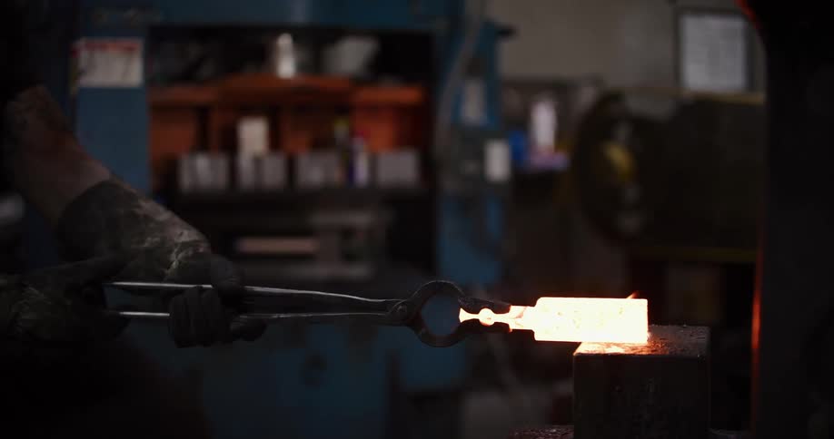 A blacksmith carefully positions a piece of red-hot, flaming metal on an anvil in a dimly lit forge