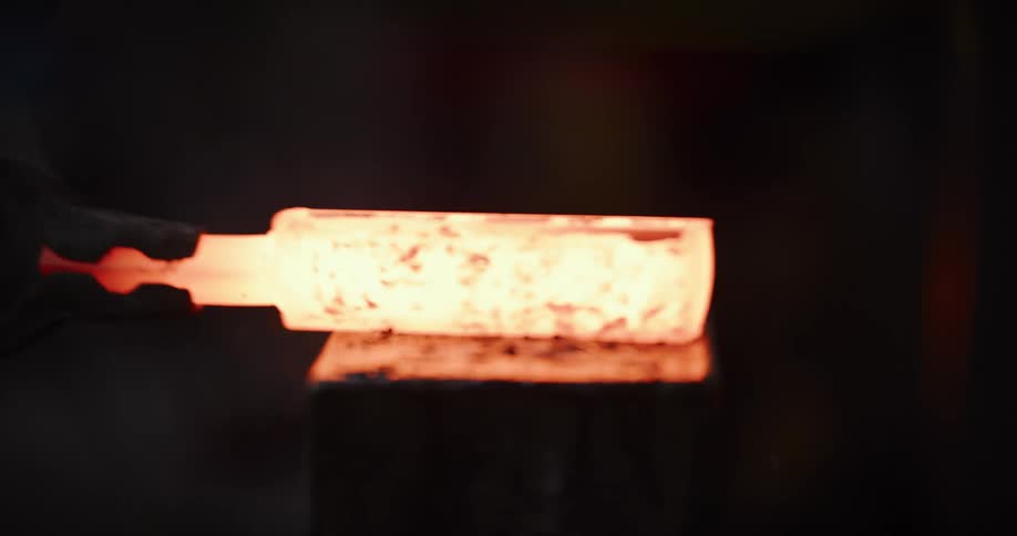 A close-up of a glowing hot metal billet held with tongs on an anvil in a dark forge