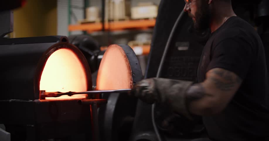 A muscular, tattooed blacksmith carefully places a piece of steel into a glowing hot forge using long tongs in his workshop