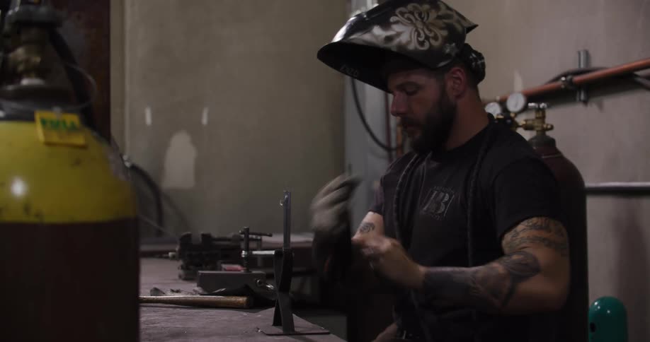 A tattooed male welder gears up with his helmet and gloves before starting to weld metal in his workshop