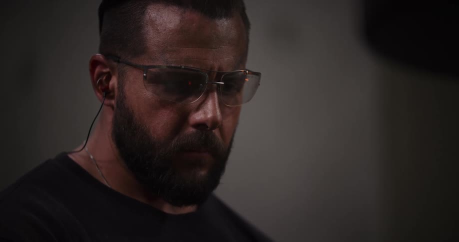 An intense close-up of a focused bearded man wearing safety glasses and ear protection in a dimly lit setting