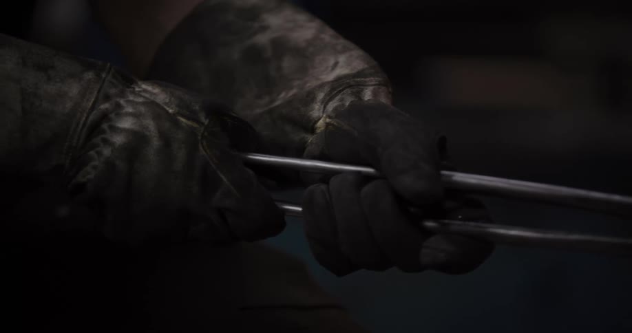 Close-up of a blacksmith's dirty gloved hands holding metal tongs in a dark workshop