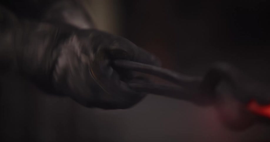 A blacksmith's gloved hand holds a piece of red-hot metal with tongs while shaping it in a dark forge