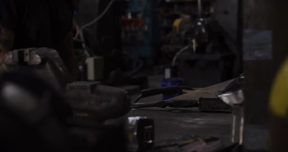 A tattooed craftsman cleans his custom welding mask and prepares metal pieces at a workbench in an industrial workshop