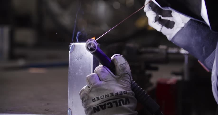 A skilled welder wearing a custom mask meticulously TIG welds a metal object in a dimly lit workshop in slow motion