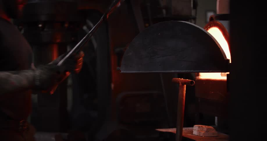 A blacksmith with tattooed arms uses tongs to place a piece of steel into a fiery furnace inside a dark workshop