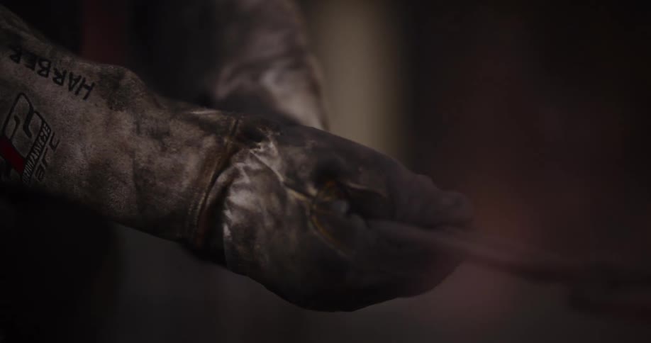 Close-up of a blacksmith's gloved hands holding a glowing red-hot piece of metal with tongs in a dark workshop