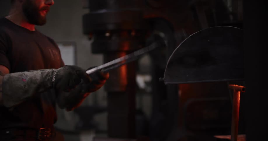 A skilled blacksmith in protective gloves inspects and handles a red-hot piece of metal with tongs in his dimly lit forge