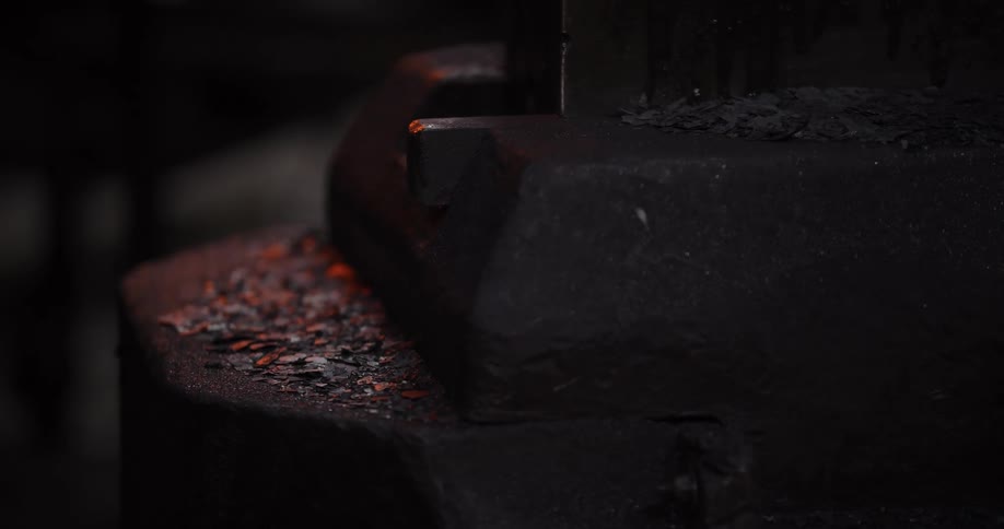 A powerful industrial press crushes glowing embers and hot metal shavings in a dark, gritty forge