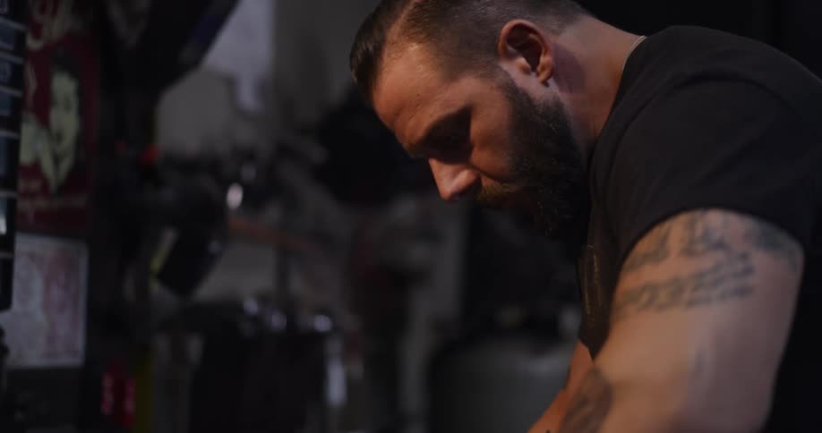 A close-up of a tattooed man with a beard concentrating on a task in a dimly lit workshop