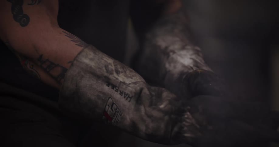 A close-up of a tattooed blacksmith's hands in worn protective gloves, using tongs to move a glowing piece of hot metal in a smoky forge