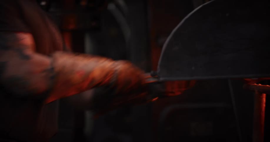 A blacksmith wearing protective gloves pulls a glowing hot piece of metal from a forge with tongs in a dark workshop
