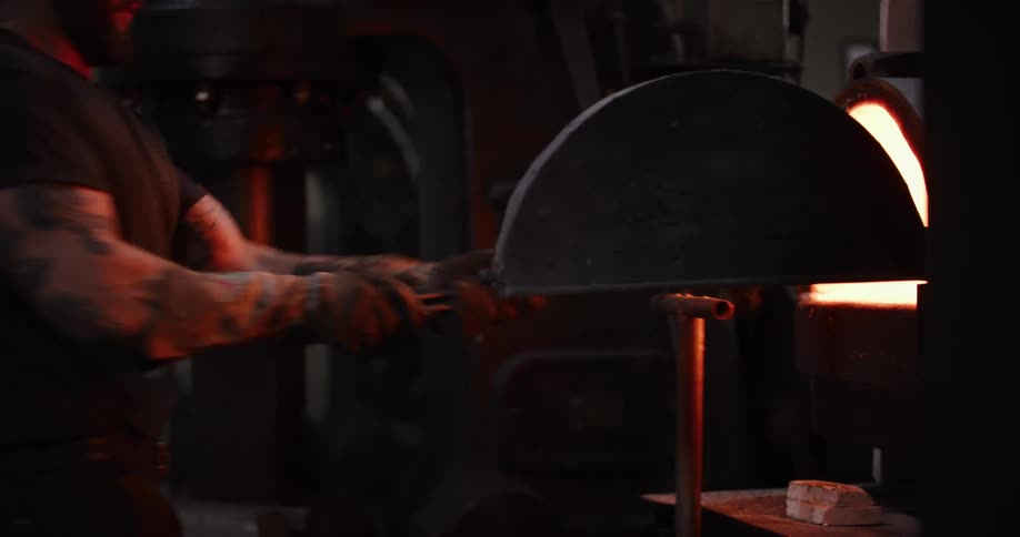 A tattooed blacksmith in a dark workshop uses tongs to pull a glowing hot piece of metal from a fiery forge