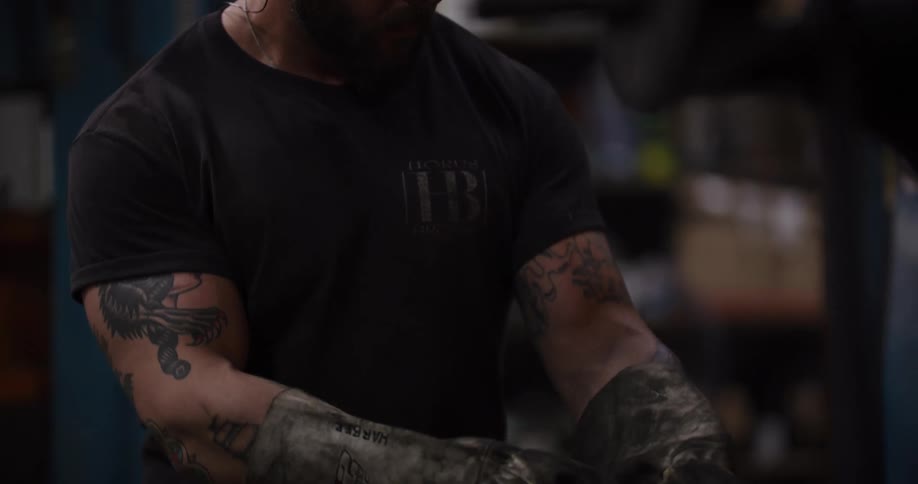 A muscular, tattooed metalworker wearing safety glasses and protective gloves intently works in his dimly lit forge