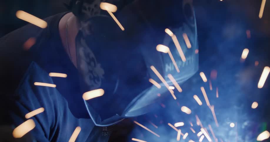 A welder in a custom protective mask meticulously works on a metal piece, creating a shower of bright sparks and smoke in dramatic slow motion