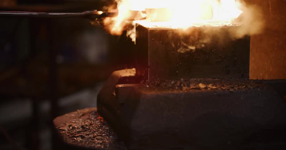 A close-up shot of a blacksmith placing a piece of red-hot steel on an anvil, causing it to smoke and burst into flames