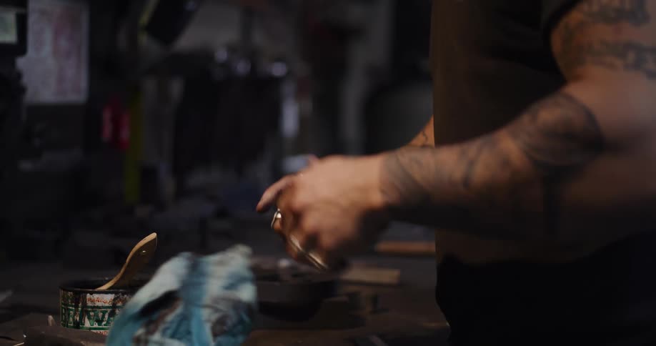 A craftsman with tattooed arms meticulously polishes and waxes a metal handle in a workshop