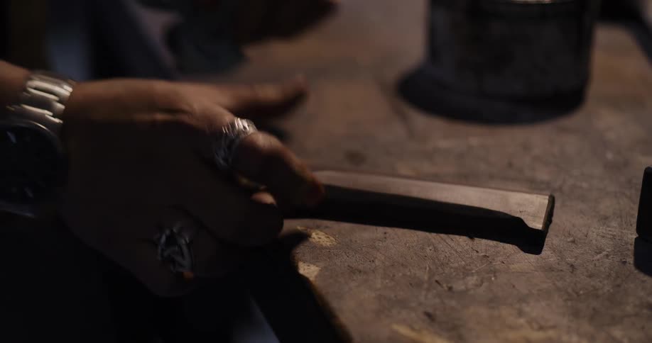 A close-up shot shows the tattooed hands of a craftsman with rings sanding a piece of metal on a rustic workbench