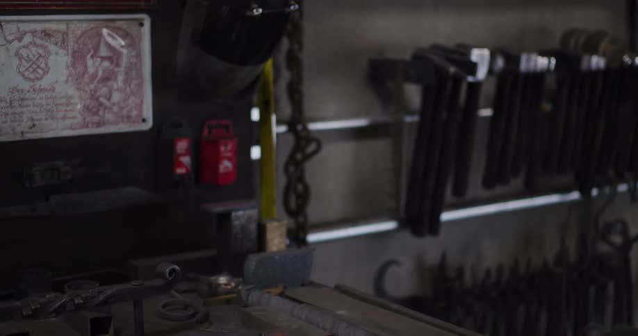Various hammers hang on a rack in a blacksmith's workshop before the camera tilts down to reveal a workbench with forged metal creations and tools