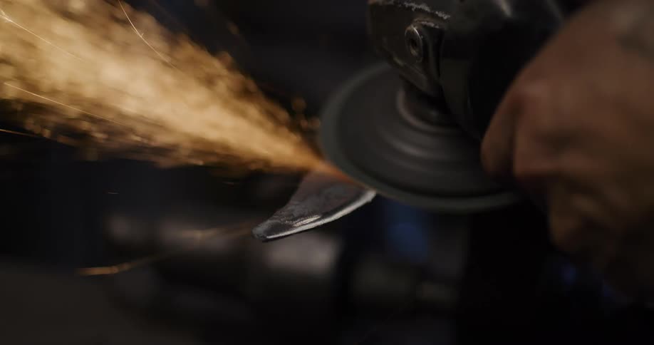A bladesmith uses an angle grinder to sharpen a metal blade, creating a shower of bright sparks in a dark workshop in slow motion