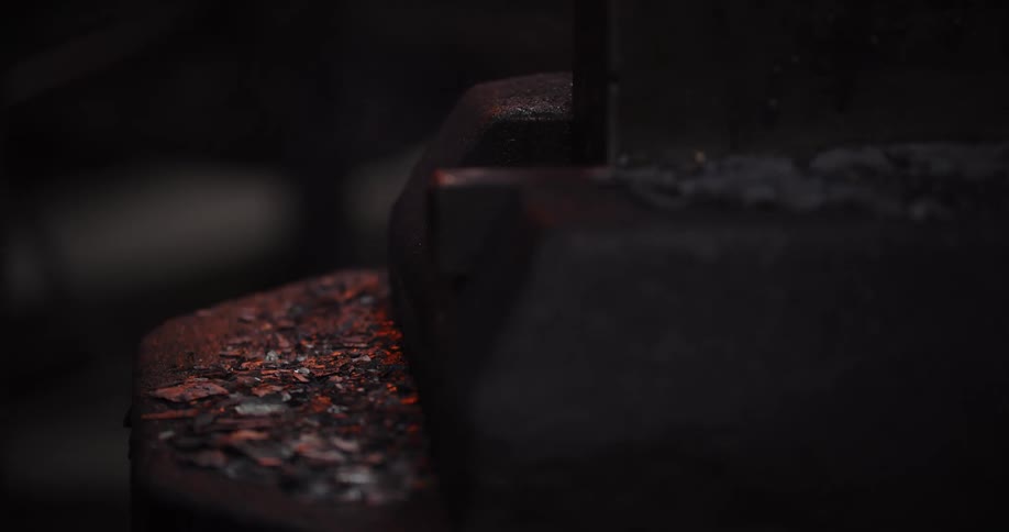 A close-up shot of a glowing hot metal ingot held by tongs cooling down on an anvil in a dark forge, its light fading away