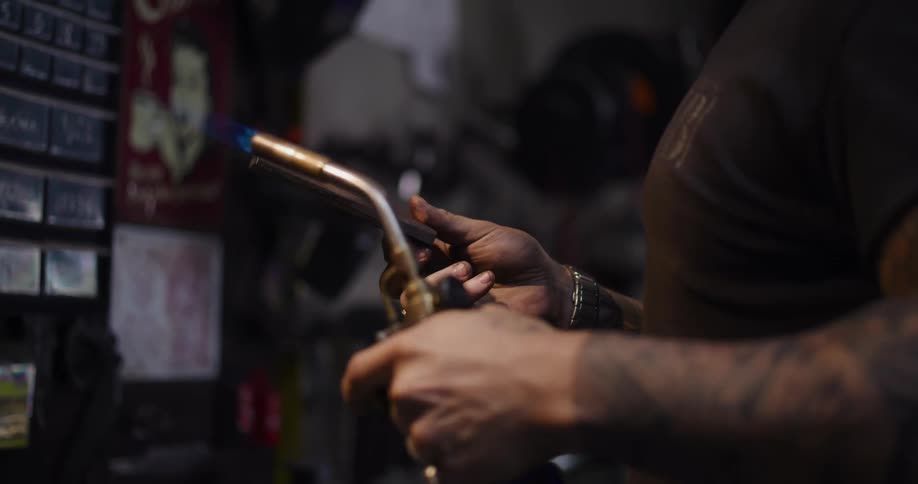 A tattooed artisan carefully heats a small piece of metal with a blowtorch in his dark workshop