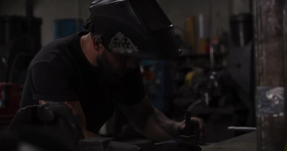 A tattooed welder puts on his gloves and mask before welding a metal piece on a workbench, creating a shower of bright sparks in a dark workshop