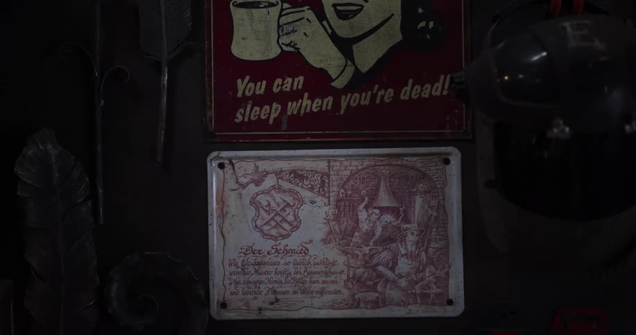 A slow tilt down reveals vintage metal signs, including a humorous coffee poster, hanging on a dark workshop wall amidst forged metal decor
