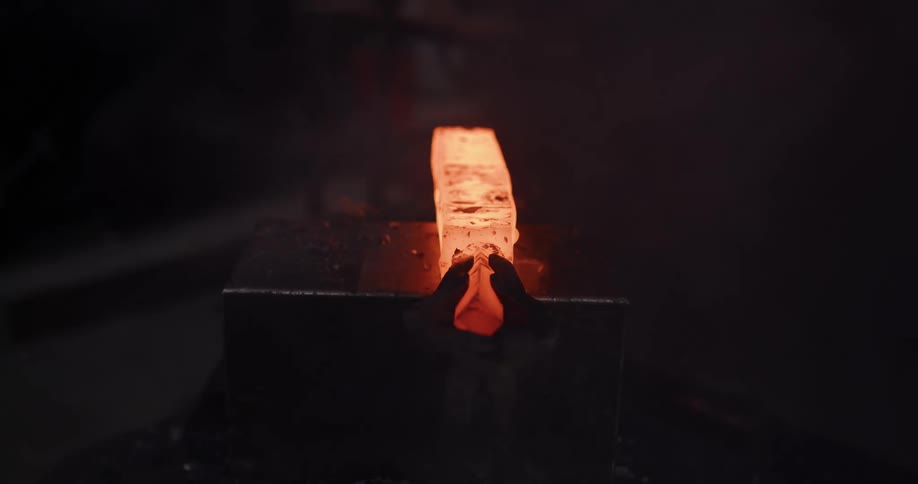 A glowing hot piece of metal is pulled from a forge with tongs and hammered into shape by an industrial power press, creating sparks and flames
