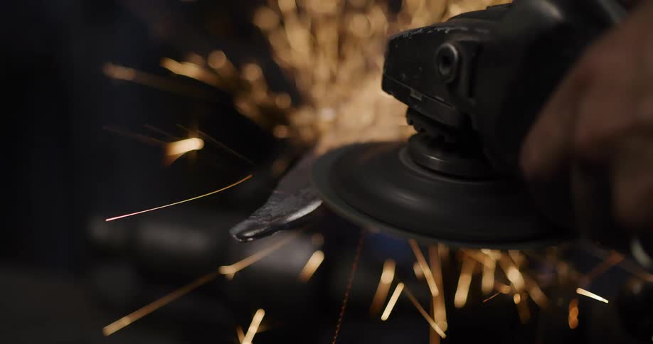 An angle grinder sends a shower of sparks flying in slow motion as it grinds a metal blade in a dark workshop