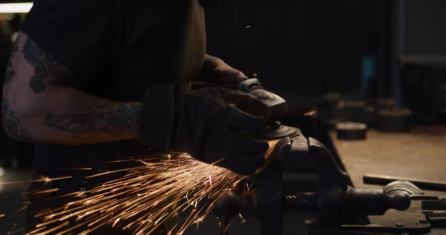 A tattooed man grinds a piece of metal in his workshop, creating a shower of sparks in slow motion