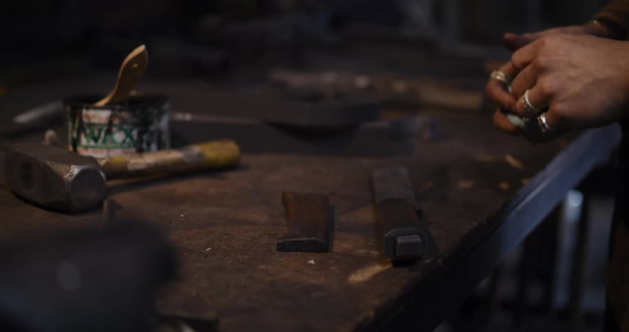 A tattooed blacksmith polishes wooden knife handle components with wax in his rustic workshop
