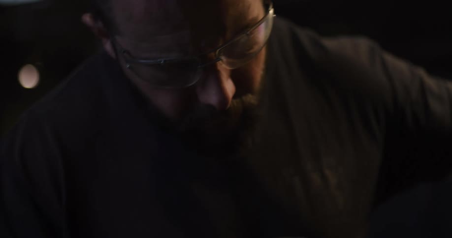 An industrial craftsman with a beard and safety glasses intensely focuses on his work, illuminated by the glow of his project