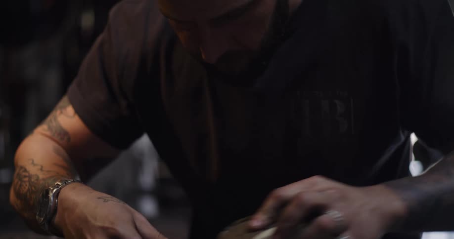 A tattooed artisan carefully sands a small wooden piece held in a vise in his dimly lit workshop