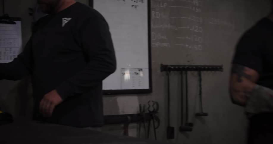 In a dimly lit industrial workshop, a metalworker carries a glowing piece of steel to the furnace while his colleague puts on protective gear