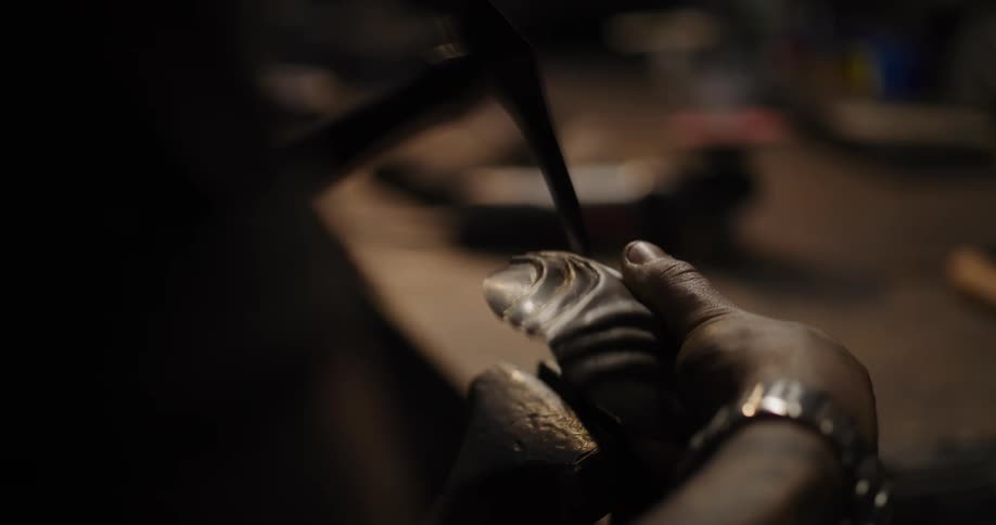A silversmith hammers an intricate pattern into a piece of metal while shaping it in a dimly lit workshop