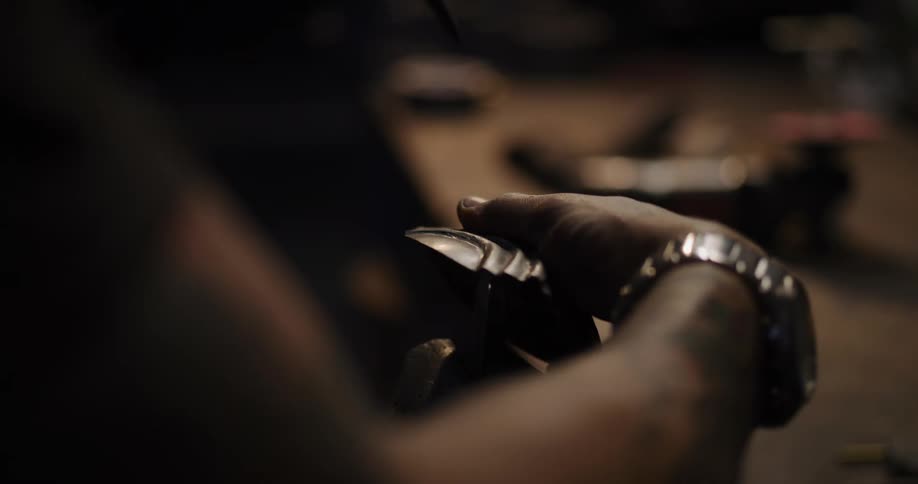 A skilled artisan with tattooed hands carefully hammers a unique piece of patterned metal in a dark workshop