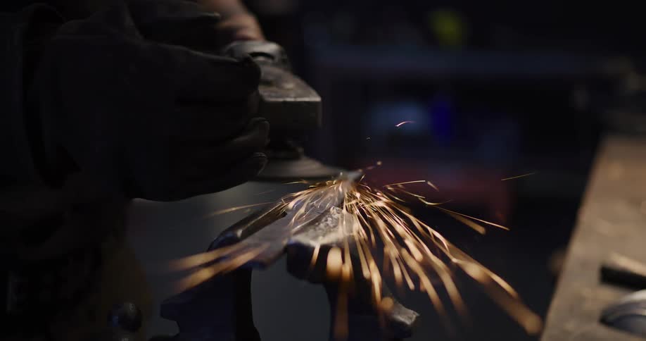 A craftsman in a dark workshop uses an angle grinder to shape a metal knife blade, sending a cascade of bright sparks flying in slow motion