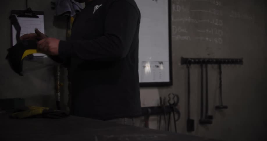 A blacksmith carries glowing hot metal through a workshop, while another worker puts on safety gear