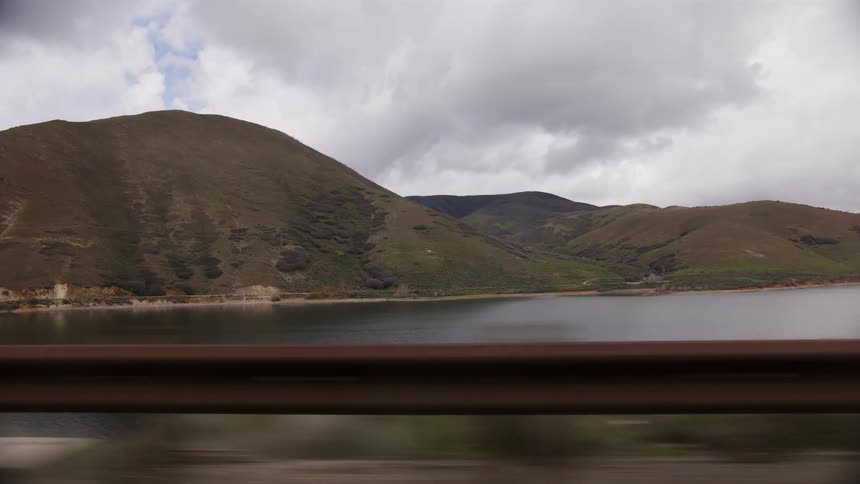 A scenic drive along a highway shows a large mountain lake and rolling green hills under a cloudy sky