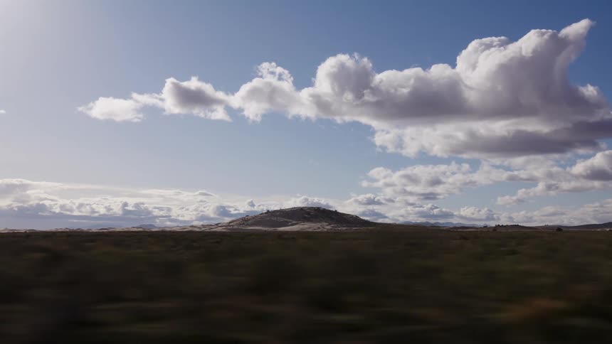 A scenic view from a moving vehicle captures a vast desert landscape and distant mountains under a beautiful cloudy sky